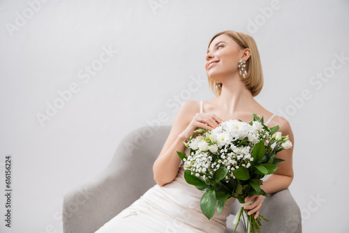special occasion, happy blonde bride in wedding dress sitting in armchair and holding bouquet on grey background, engagement ring, white flowers, bridal accessories, happiness, feminine