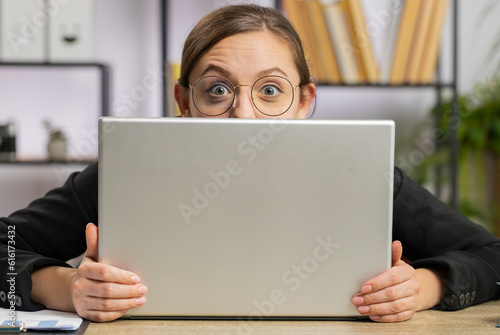 Confident business woman hiding behind laptop computer, looking at camera, spying his colleagues working, peeping. Professional freelancer girl looking from behind computer with cunning eyes glance