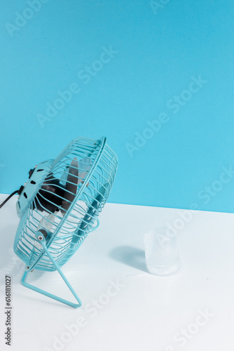  Hot temperature concept with a little blue fan cooling an ice cube on white and blue background. Vertical view