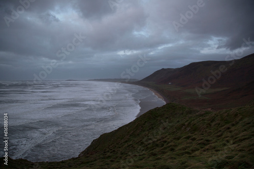Rhossili Bay at dusk during a winter's storm with dramatic skies, wind and rain. Rhossili Downs are covered with bracken and waves are pounding the beach at high tide.