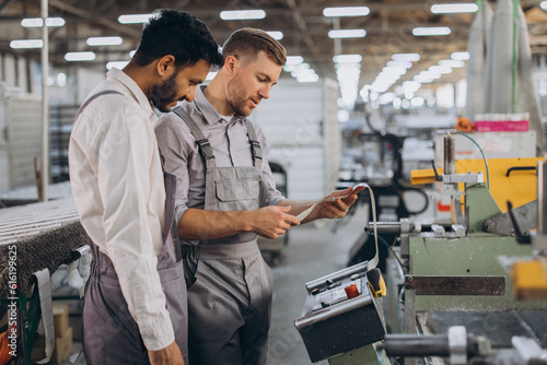 A male inspector or operator of a workshop for the production of aluminum and plastic wreaths trains an intern. International team of men working together near a machine in a factory.