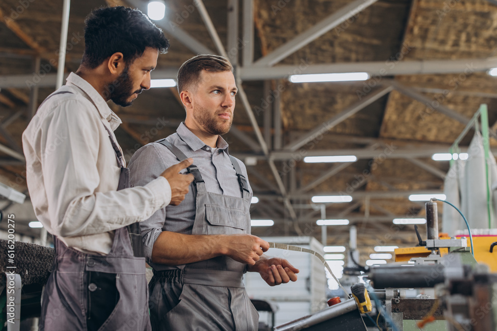 A male inspector or operator of a workshop for the production of aluminum and plastic wreaths trains an intern. International team of men working together near a machine in a factory.