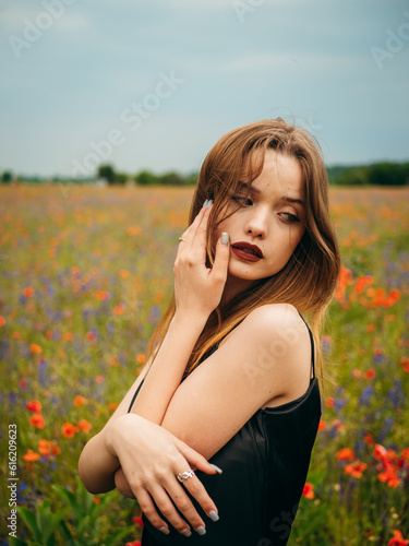 Portrait of a beautiful young girl in a black evening dress posing against a poppy field on a cloudy summer day. Portrait of a female model outdoors. Rainy weather. Gray clouds. Vertical shot.