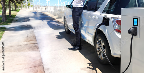 charging electric vinhicle on ev station ner public park and  wind turbine and man near car breesing clear air