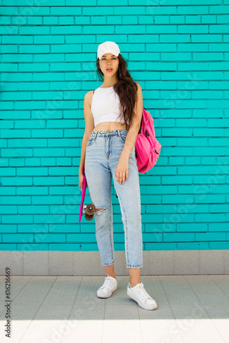 teen girl skater. young fit brunette girl in white cap and jeans stands casual with pink skate in hand near blue brick wall background and looks straight with cute smile. lifestyle concept, free space