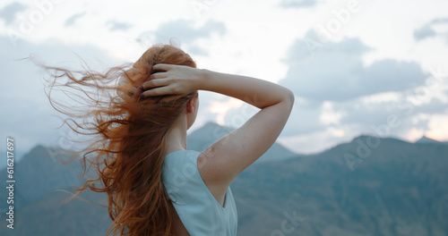 Back view of beautiful cheerful redhead girl with flying curly hair and clothing in light dress looking at mountains and clouding sky. Unity with Nature Concept. Peace and tranquility. Copy space.