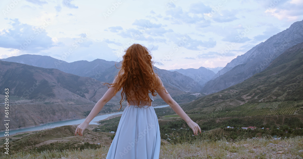 Beautiful carefree girl with red hair wearing white dress standing against the wind, looking at mountains and raising her hands - freedom, adventure, inspiration 