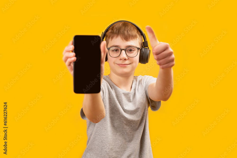 School boy in headphones and glasses holds smartphone with blank screen and shows thumbs up. Apps for listening music or learning. Copy space School boy in headphones and glasses holds smartphone with blank screen and shows thumbs up. Apps for listening music or learning. Copy space