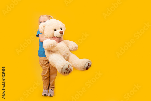 Little blond girl hugs large toy bear. Studio portrait of child with toy on bright yellow background. Copy space
