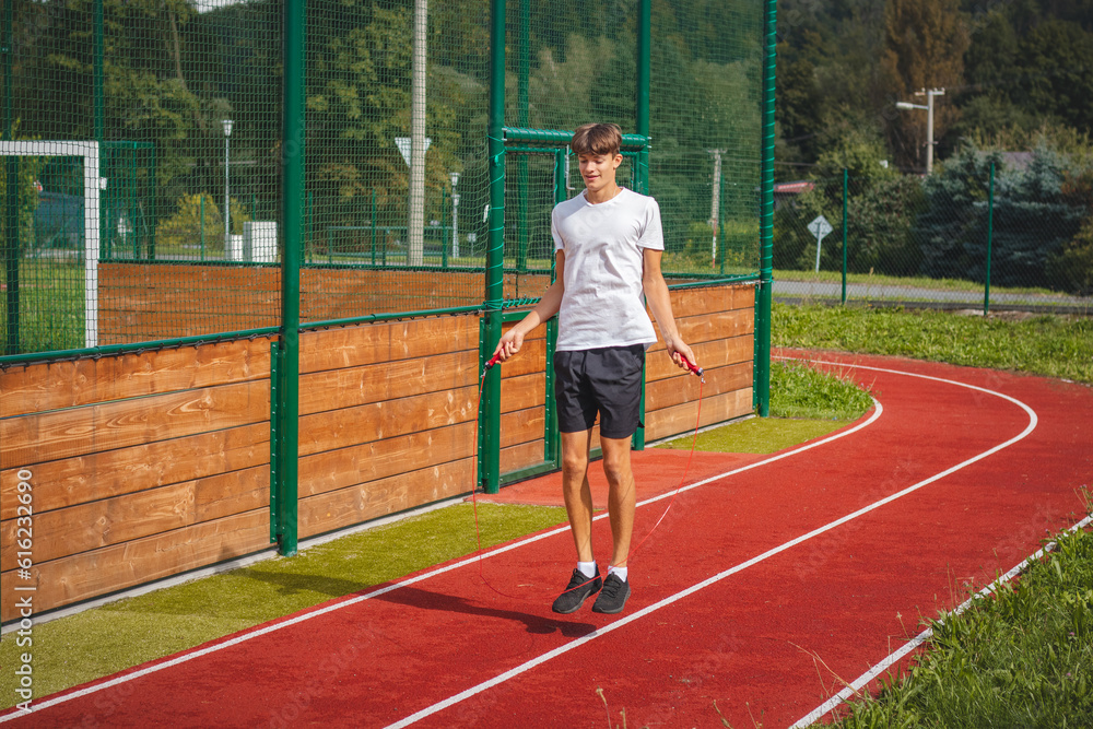 Brown-haired boy with an athletic figure wearing a white T-shirt and black shorts is jumping rope on an athletic oval. Training to improve jumping, coordination and endurance strength