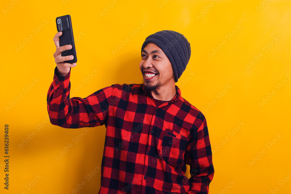 Asian man in a plaid shirt and beanie hat holding a mobile phone isolated on yellow background. Asian man in a plaid shirt and beanie hat holding a mobile phone isolated on yellow background.