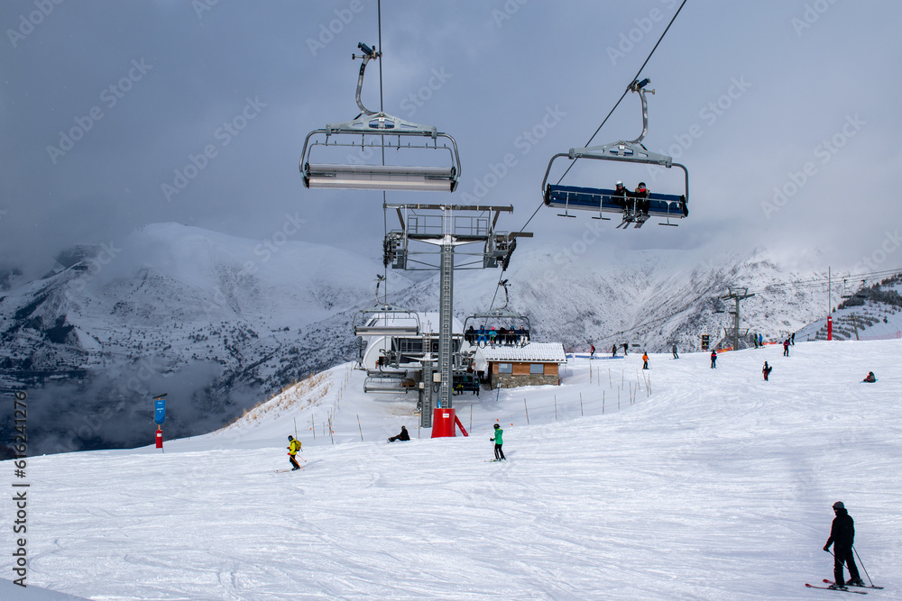 On top of a snow-covered ski resort with skiers and ski lifts passing in the foreground and snowy mountains in the background, Les Deux Alpes, France, January 2023
