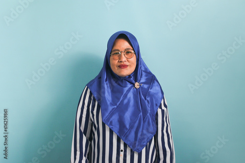 Portrait of a middle-aged Asian woman in a blue hijab and striped shirt, wearing eyeglasses and looking at the camera. She is isolated over a blue background