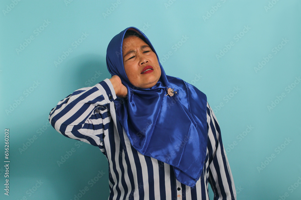 Asian woman in blue hijab massages neck/shoulders, relieving muscle pain from overwork, incorrect posture, against blue background. Asian woman in blue hijab massages neck/shoulders, relieving muscle pain from overwork, incorrect posture, against blue background.