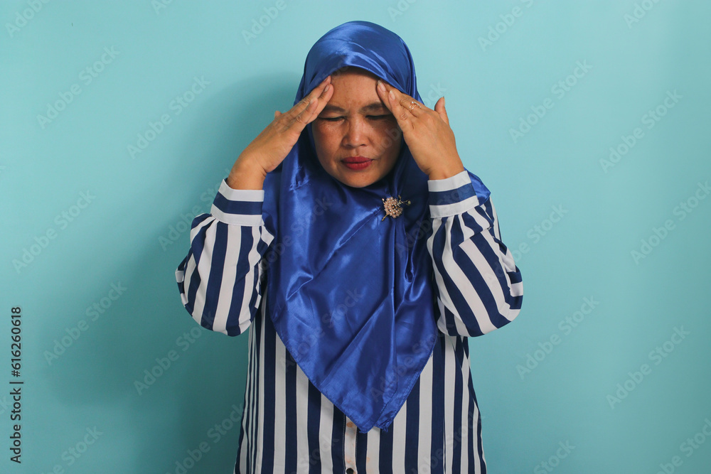 Portrait of a middle-aged Asian woman, wearing a blue hijab and striped shirt, shows her suffering from a headache, migraine, or illness, standing against a blue background