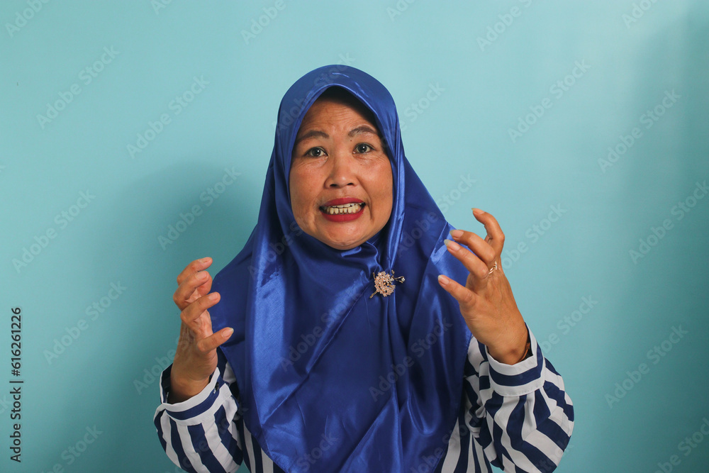 A furious middle-aged Asian woman, wearing a blue hijab and a striped shirt, raises her hands and looks at the camera while standing against a blue background A furious middle-aged Asian woman, wearing a blue hijab and a striped shirt, raises her hands and looks at the camera while standing against a blue background