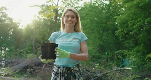 Portrait of volunteer woman holding plant in forest. Young woman dressed in blue volunteer t-shirt holds in hands little plant sprout. Voluntary free work assistance help, charity grace concept. photo