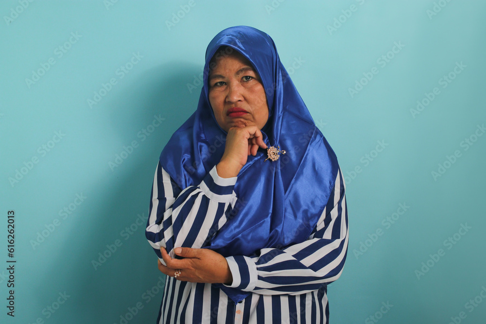A pensive middle-aged Asian woman in a blue hijab and striped shirt touches her chin, looking confused and worried while standing against a blue background A pensive middle-aged Asian woman in a blue hijab and striped shirt touches her chin, looking confused and worried while standing against a blue background