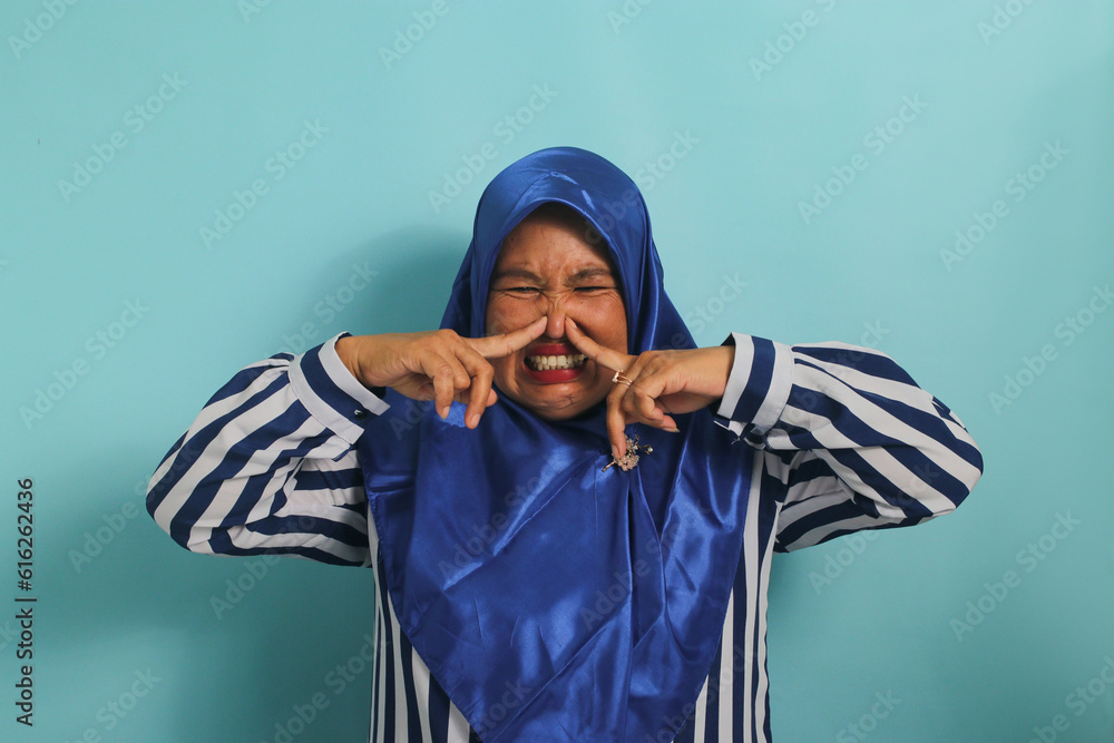 A middle-aged Asian woman, in a blue hijab, holds her breath and pinches her nose with her fingers, reacting to a stinky and disgusting, intolerable bad smell, while standing against a blue background - obrazy, fototapety, plakaty 