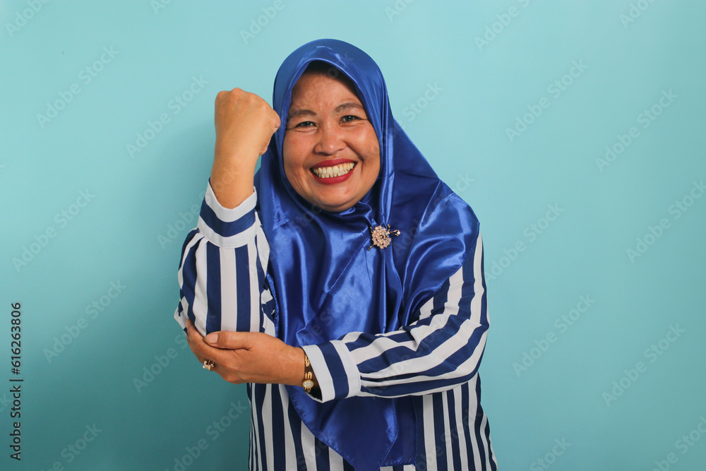 A smiling middle-aged Asian woman, in a blue hijab and striped shirt, makes a strong gesture by lifting her arms and flexing her muscles towards the camera, standing against a blue background A smiling middle-aged Asian woman, in a blue hijab and striped shirt, makes a strong gesture by lifting her arms and flexing her muscles towards the camera, standing against a blue background