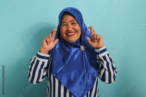 An excited middle-aged Asian woman, wearing a blue hijab, is hopeful and optimistic, crossing her fingers for good luck, making a wish, and smiling while standing against a blue background