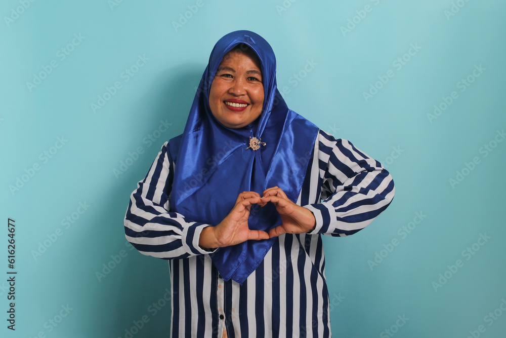A smiling middle-aged Asian woman, in a blue hijab and striped shirt, makes a heart gesture with her hands, radiating happiness and care, isolated on blue background A smiling middle-aged Asian woman, in a blue hijab and striped shirt, makes a heart gesture with her hands, radiating happiness and care, isolated on blue background