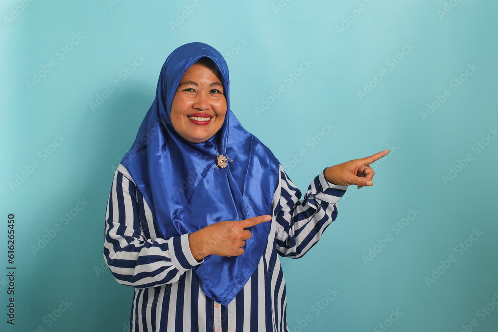 An enthusiastic middle-aged Asian woman, wearing a blue hijab and striped shirt, points to the right, emphasizing the copyspace. Isolated over blue background. An enthusiastic middle-aged Asian woman, wearing a blue hijab and striped shirt, points to the right, emphasizing the copyspace. Isolated over blue background.