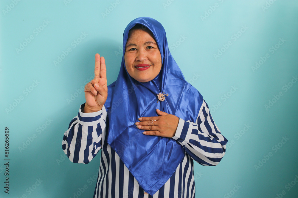 A Confident middle-aged Asian woman in a blue hijab and striped shirt is making a swearing or promise gesture while looking at the camera with hand on chest. She is isolated over a blue background.