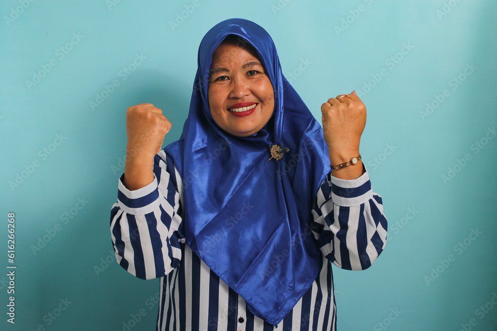 An excited middle-aged Asian woman in a blue hijab and striped shirt shows a strong gesture by lifting her arms and flexing her muscles, smiling proudly at the camera, isolated on blue background. An excited middle-aged Asian woman in a blue hijab and striped shirt shows a strong gesture by lifting her arms and flexing her muscles, smiling proudly at the camera, isolated on blue background.