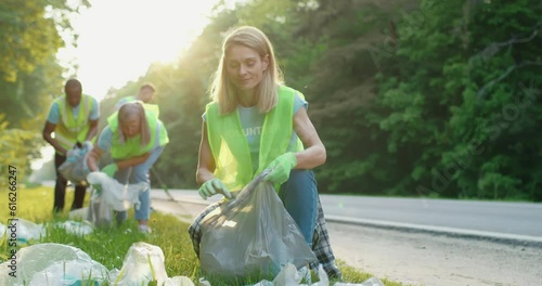 Portrait of beautiful girl with small group of volunteers on background with gloves and garbage bags cleaning up area near road. Environment preservation and ecology concept.