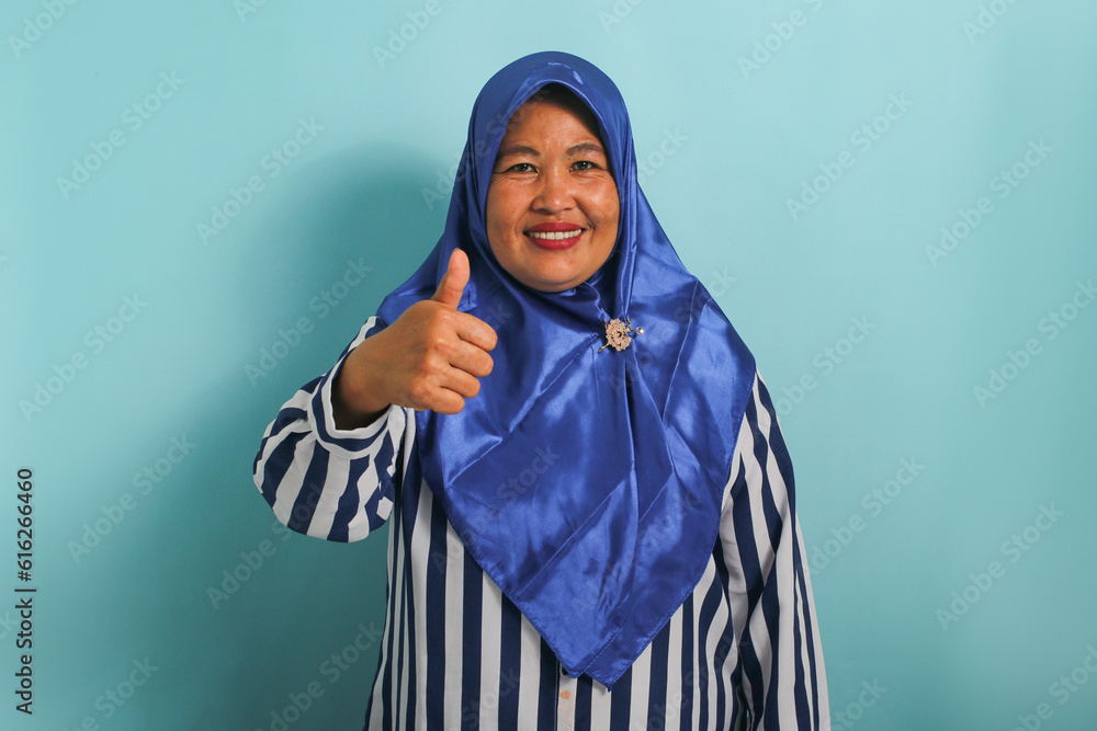 An excited middle-aged Asian woman in a blue hijab and striped shirt shows a thumbs-up and an okay sign, symbolizing success and expressing approval, while standing against a blue background. An excited middle-aged Asian woman in a blue hijab and striped shirt shows a thumbs-up and an okay sign, symbolizing success and expressing approval, while standing against a blue background.