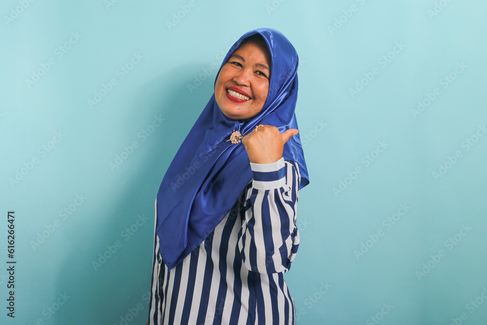 An excited middle-aged Asian woman in a blue hijab and striped shirt points to the left with her thumb, highlighting the copyspace or empty space, isolated over blue background. An excited middle-aged Asian woman in a blue hijab and striped shirt points to the left with her thumb, highlighting the copyspace or empty space, isolated over blue background.