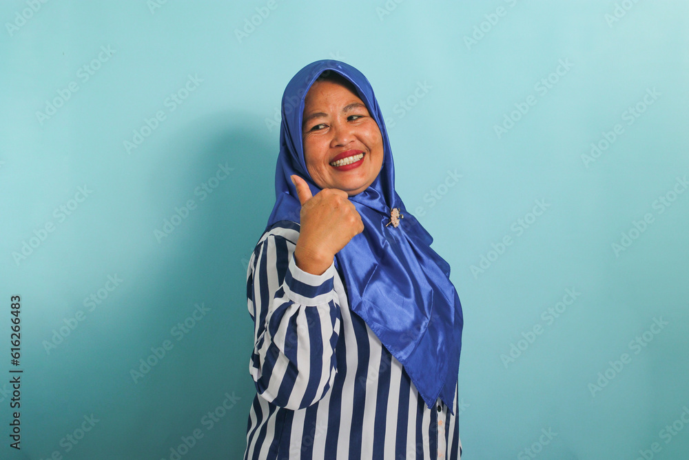 An excited middle-aged Asian woman in a blue hijab and striped shirt points to the left with her thumb, highlighting the copyspace or empty space, isolated over blue background. An excited middle-aged Asian woman in a blue hijab and striped shirt points to the left with her thumb, highlighting the copyspace or empty space, isolated over blue background.