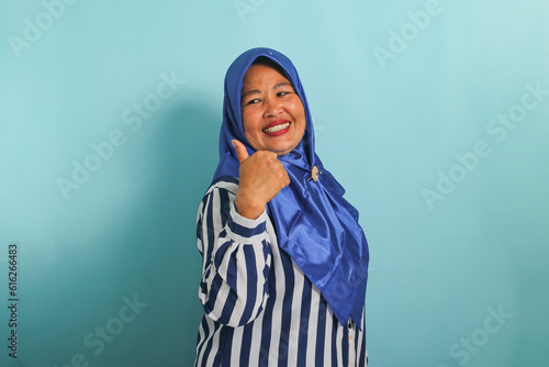 An excited middle-aged Asian woman in a blue hijab and striped shirt points to the left with her thumb, highlighting the copyspace or empty space, isolated over blue background.