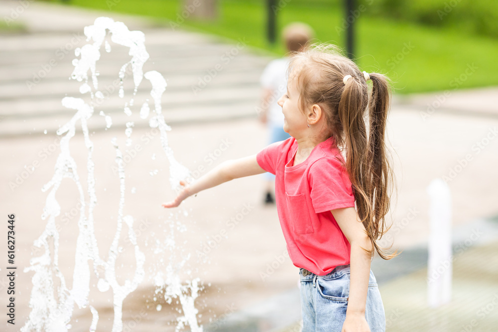 Naklejka premium A happy little girl has fun near the city fountain. The child has a rest near a fountain during a heat wave.