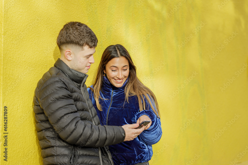Young couple look at a mobile phone with a yellow wall in the background with copy space.
