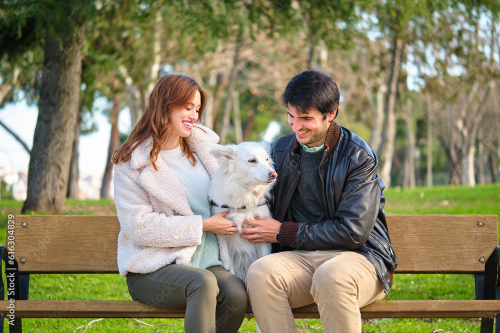 Fototapeta premium Young caucasian couple petting their dog sitting on a bench in a park. Family.