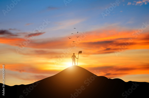 Silhouette of businessman holding flag on the top of mountain at sunrise and bird flying over the sky. It is symbol of leadership successful achievement with goal and objective target of company.