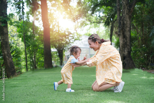 Baby girl is doing her first steps with mother help. Cute little girl learns to walk with her mom helping her in the sunny garden outdoors. Happy family moment concept. © Bordinthorn
