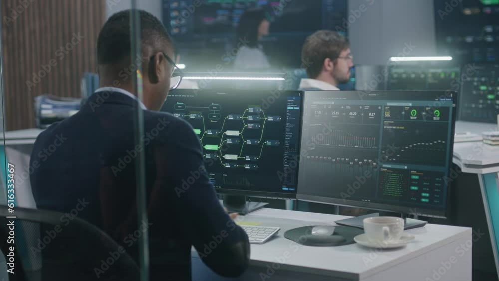 African American big data scientist works at computer in monitoring room. Multiracial IT engineers analyze charts at background. Multiple big screens on the wall with displayed blockchain network map. African American big data scientist works at computer in monitoring room. Multiracial IT engineers analyze charts at background. Multiple big screens on the wall with displayed blockchain network map.