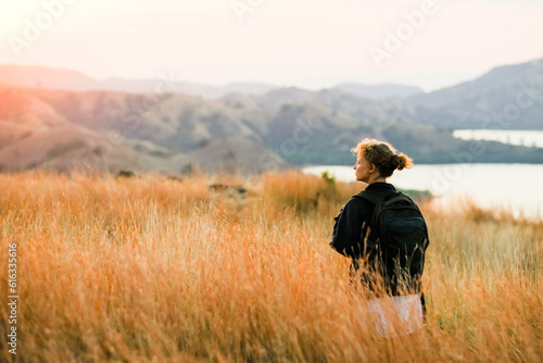 A young girl, blonde, with a backpack, stands on top of a mountain, rear view, overlooking a beautiful panorama of the islands and bays in Indonesia.