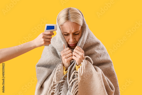 Ill mature woman measuring temperature on yellow background