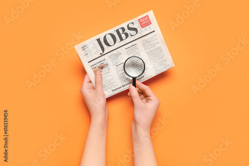 Female hands with mini magnifier and newspaper with headline JOBS on orange background. Search concept