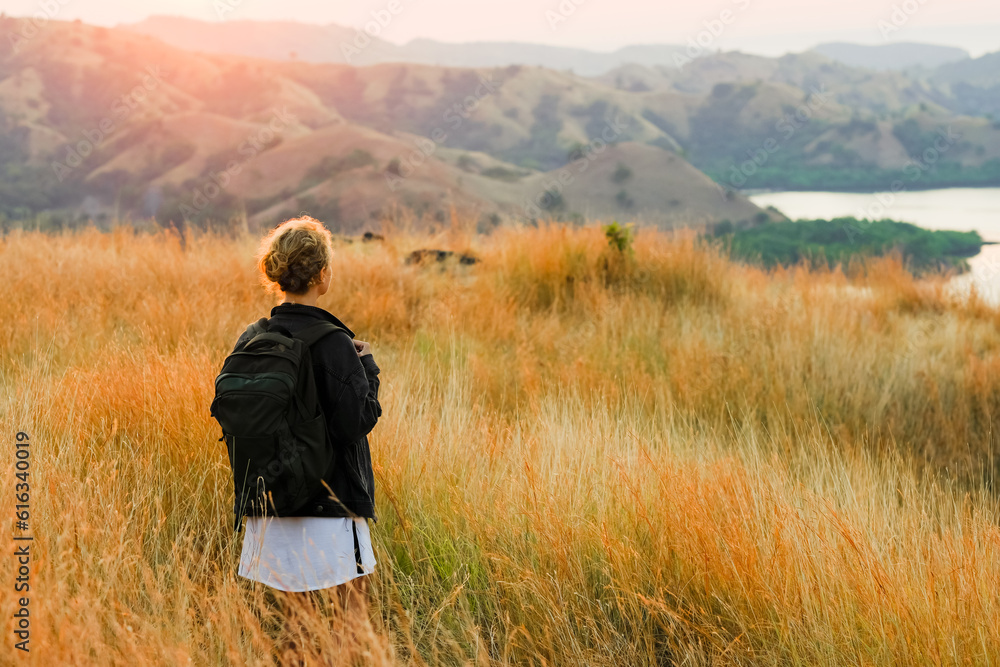 A young girl, blonde, with a backpack, stands on top of a mountain, rear view, overlooking a beautiful panorama of the islands and bays in Indonesia.
