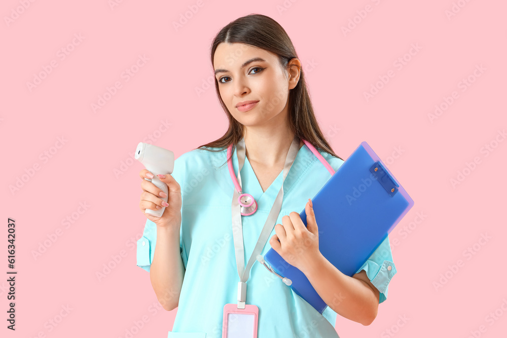 Female medical intern with infrared thermometer and clipboard on pink background Female medical intern with infrared thermometer and clipboard on pink background