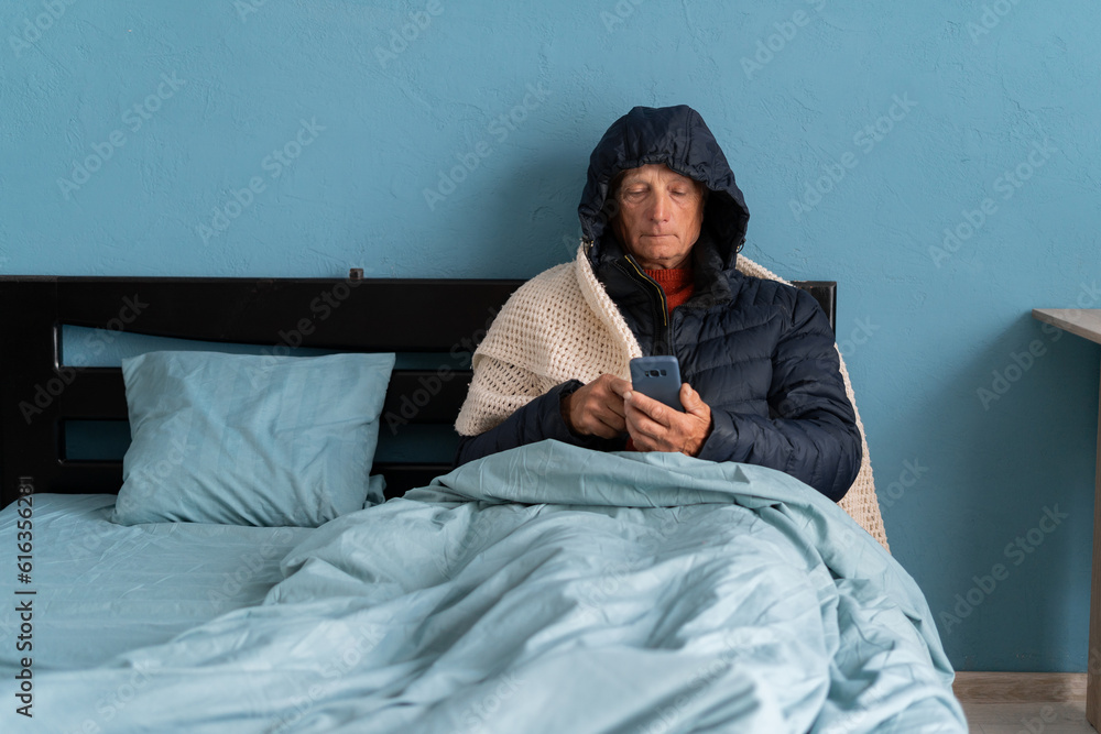 Sick senior man with phone, feel cold covered with blanket sit in bed, shiver freezing warming at home, no central heating, problem and flu concept Sick senior man with phone, feel cold covered with blanket sit in bed, shiver freezing warming at home, no central heating, problem and flu concept