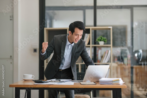 A cheerful Asian businessman in a formal business suit is raising his hands, looking at his laptop screen, and celebrating his success in the office. project approved, getting job promotion