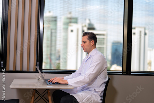 Doctor in a white coat sitting working on laptop with a coffee cup on a table in the pantry room