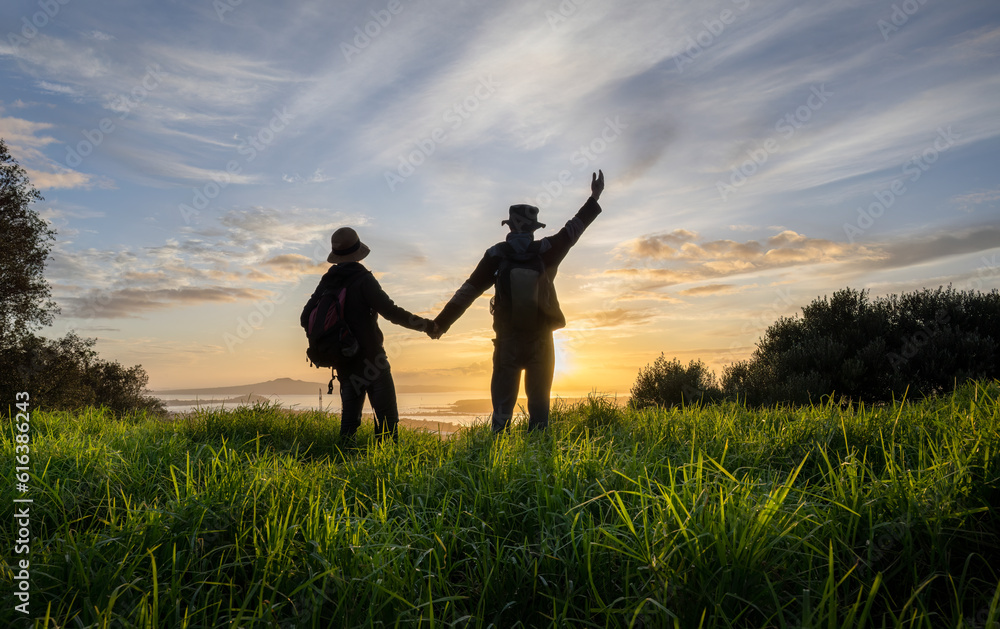 Couple holding hands and standing on Mt Eden summit at sunrise. Rangitoto Island in the distance. Auckland.