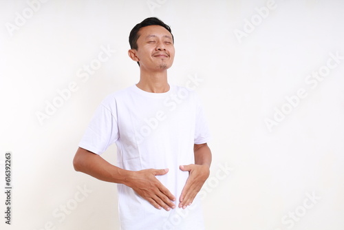 Asian funny man standing while holding his tummy. He feels full. Isolated on white background